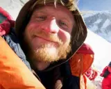Uśmiechnięty alpinista w ciepłej kurtce, z brodą, na tle ośnieżonych gór. Może to być jego lek na chorobę wysokościową.