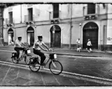 Due agenti in bicicletta pattugliano una strada italiana. Sul marciapiede, una coppia passeggia con i loro cani. Street photography Italia.