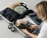 Kobieta pakuje walizkę podręczną: ubrania, buty, torebka, okulary, perfumy.