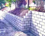 Un muro de contención de bloques de hormigón se está construyendo en un jardín, con tierra y vegetación detrás.