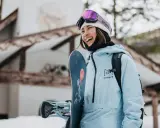 Uśmiechnięta snowboardzistka w jasnoniebieskim anoraku i goglach Anon. Gotowa na stok, trzyma deskę Burton.