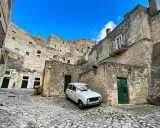 Un'auto d'epoca bianca parcheggiata tra le antiche case scavate nella roccia dei Sassi di Matera, sotto un cielo azzurro.