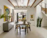 Comedor moderno en una casa entre medianeras, con vigas de madera en el techo, mesa de madera, sillas negras y una escalera de madera.