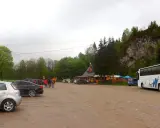 Parking przy Wąwozie Homole. Samochody, autokar i budynek zadaszony, otoczony drzewami, u podnóża skalnego zbocza.