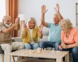 Radosna terapia zajęciowa dla seniorów: czworo uśmiechniętych osób gra w Jengę, unosząc ręce w geście zwycięstwa lub ekscytacji.