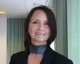 En kvinna med glasögon på huvudet ler. Hon bär en svart kavaj och en grå scarf.