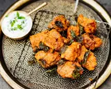 Knusprige chicken pakora auf einem Gitter, garniert mit frischen Kräutern, daneben eine Schale Joghurt-Dip.
