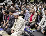 Männer in traditioneller Kleidung knien auf Gebetsteppichen. Ein Mann in einem weißen Gewand und Turban leitet das Gebet, ein Zeichen der berberischen Religion.