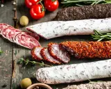 Assortiment de charcuteries, dont une spécialité des Pyrénées enrobée d'épices, sur fond de bois rustique avec tomates et herbes.