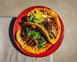 Zwei saftige Birria Tacos auf einem roten Teller, garniert mit Koriander und Limettenspalten.