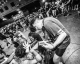 Energetyczny koncert w klubie. Czarno białe zdj. z tłumem szalejącym pod sceną, gdzie gitarzysta pochyla się nad instrumentem.