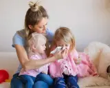 Mama wyciera nos chorej córce, podczas gdy młodsza siostra patrzy. Czy szczepienie mogłoby zapobiec katarowi?