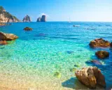 Acque cristalline e rocce a picco sul mare bello in Campania, con i Faraglioni di Capri all'orizzonte.