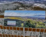Widok z wieży widokowej na ośnieżony las i panoramę gór z tablicą informacyjną.