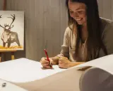 En kvinna ler medan hon ritar på ett stort papper. I bakgrunden syns en oljemålning av en ren. Hon deltar i en oljemålning kurs.