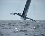Un maxi trimaran fend les vagues, ses coques latérales hors de l'eau, propulsé par une voile immense. L'équipage est visible sur le pont.
