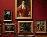Obrazy w bogatym, malarskim stylu zdobią czerwoną, wzorzystą ścianę. Portret mężczyzny w stroju z epoki, bukiet kwiatów i sceny rodzajowe tworzą galerię.