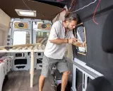 Mężczyzna buduje kamper z osobówki, montując okno. Widać drewniane elementy łóżka i instalację elektryczną.