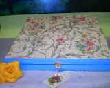 Caja azul con tapa decorada con papel decoupage floral. Un detalle de rosa amarilla y etiqueta floral completan la escena.