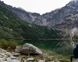 Osoba siedzi nad jeziorem w górach, podziwiając skaliste szczyty. To jedna z wielu pięknych bukowina tatrzańska atrakcji.