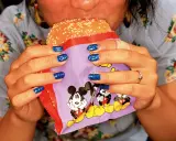 Mani con smalto blu intenso tengono un hamburger in un involucro con Topolino, in uno stile tipico di Martin Parr.
