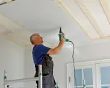 Man spacklar med remsa i taket, skruvar gipsskivor på träreglar.