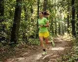 Frau joggt durch einen sonnendurchfluteten Wald. Sie trägt ein grünes Shirt und gelbe Shorts. Das zeigt, was bringt joggen: Natur, Bewegung und Wohlbefinden.