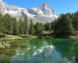 Un gruppo di escursionisti ammira il riflesso del Cervino nel lago blu di Cervinia, circondato da pini e montagne innevate.