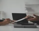 Dwie osoby omawiają dokument, jedna wskazuje piórem na klauzulę CV. Obok leży laptop i kalkulator.