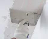 Ręka w rękawicy nakłada zaprawę na styropian przy suficie. Proces pokazujący, jak przykleić styropian do sufitu.
