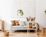 Un salón moderno con un sofá gris, cojines étnicos y una manta mostaza. La decoración se completa con una planta colgante y un jarrón con hierbas secas.