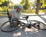 Mężczyzna w kasku siedzi na drodze obok przewróconego roweru, trzymając się za bolącą nogę po wypadku rowerowym.