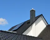 Nowoczesny komin na dachu z czarnej dachówki, z panelem słonecznym. Jaka średnica komina do pieca na drewno?