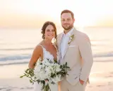 Groom in a tan suit, perfect for what to wear to beach wedding male, smiles with bride holding bouquet at sunset.