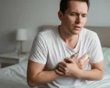 Mann hält sich die Brust, leidet unter Brustmuskel Schmerzen. Er sitzt auf einem Bett, sein Gesichtsausdruck ist besorgt.