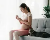 Schwangere Frau isst Joghurt, Katze liegt daneben. Achtung bei Toxoplasmose Symptome Schwangerschaft.