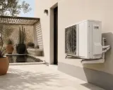 Unité extérieure d'une pompe à chaleur air air ou air eau, fixée sur un mur de maison, avec une piscine et des plantes en arrière-plan.