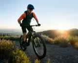 Kolarz w kasku i okularach jedzie rowerem po górskiej ścieżce. Słońce zachodzi za wzgórzami, tworząc piękne światło. To idealny moment na maraton MTB.