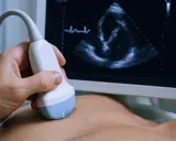 Dłoń trzyma sondę USG, która bada echo serca co wykrywa jego pracę. Na ekranie widać obraz serca i zapis EKG.