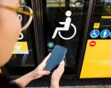Osoba trzyma telefon przed drzwiami autobusu z symbolem wózka inwalidzkiego. To ułatwia podróżowanie, gdy masz legitymację osoby niepełnosprawnej.