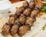Kofta kebab z bakłażanem na patyku, podany na płaskim chlebie z sosem.