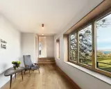 Interior moderno con suelo de madera, sillón gris y gran ventanal. El marco de madera de la ventana, que parece recién restaurado, enmarca un paisaje exterior.