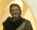 Kurt Russell som Ego i filmer med kurt russell, med grått hår och skägg, klädd i en mörk sjal och rustning mot en gul bakgrund.
