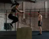 Frau springt auf eine Box, um ihre Kondition aufzubauen. Mann beobachtet sie im Fitnessstudio.