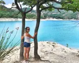 Donna sorridente abbraccia un albero con un lago turchese sullo sfondo. Una perfetta gita fuori porta nelle Marche.