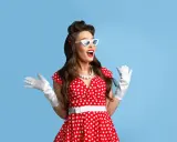 Kobieta w czerwonej sukience w białe grochy, białych rękawiczkach i okularach w stylu pin-up girl stylizacja, z uniesionymi rękami.