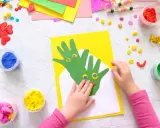 Kreatives Basteln mit Kleinkindern: Hände werden zu grünen Drachen auf gelbem Papier, umgeben von bunter Knete und Bastelmaterialien.