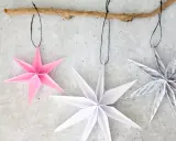 Drei Papiersterne, die Kinder falten könnten, hängen an einem Ast. Einer ist rosa, die anderen weiß und mit Noten bedruckt.