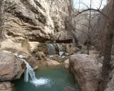 Cascada y río en un cañón rocoso, con hielo en las rocas. Un puente de madera cruza el río, evocando las pasarelas de Valloré.
