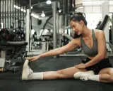 Junge Frau dehnt sich im Fitnessstudio, um ihre **Beweglichkeit zu verbessern**. Sie sitzt auf dem Boden und streckt ein Bein nach vorne.